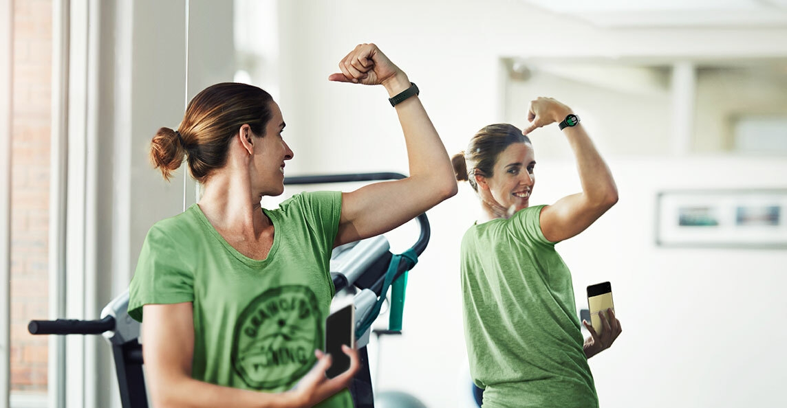 Une femme regarde le miroir et contracte ses muscles. Elle porte une Galaxy Watch 4 au poignet et un Galaxy Z Flip 3 5G dans sa main droite.